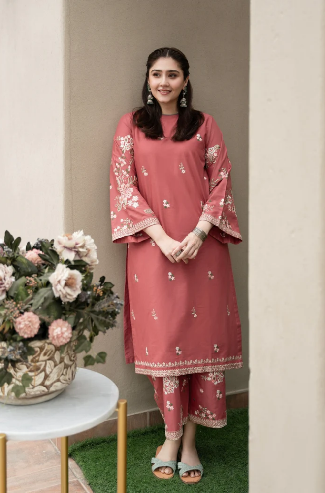 A woman wearing a pink kurta and pants stands beside a potted plant, smiling gently in a bright, natural setting.
